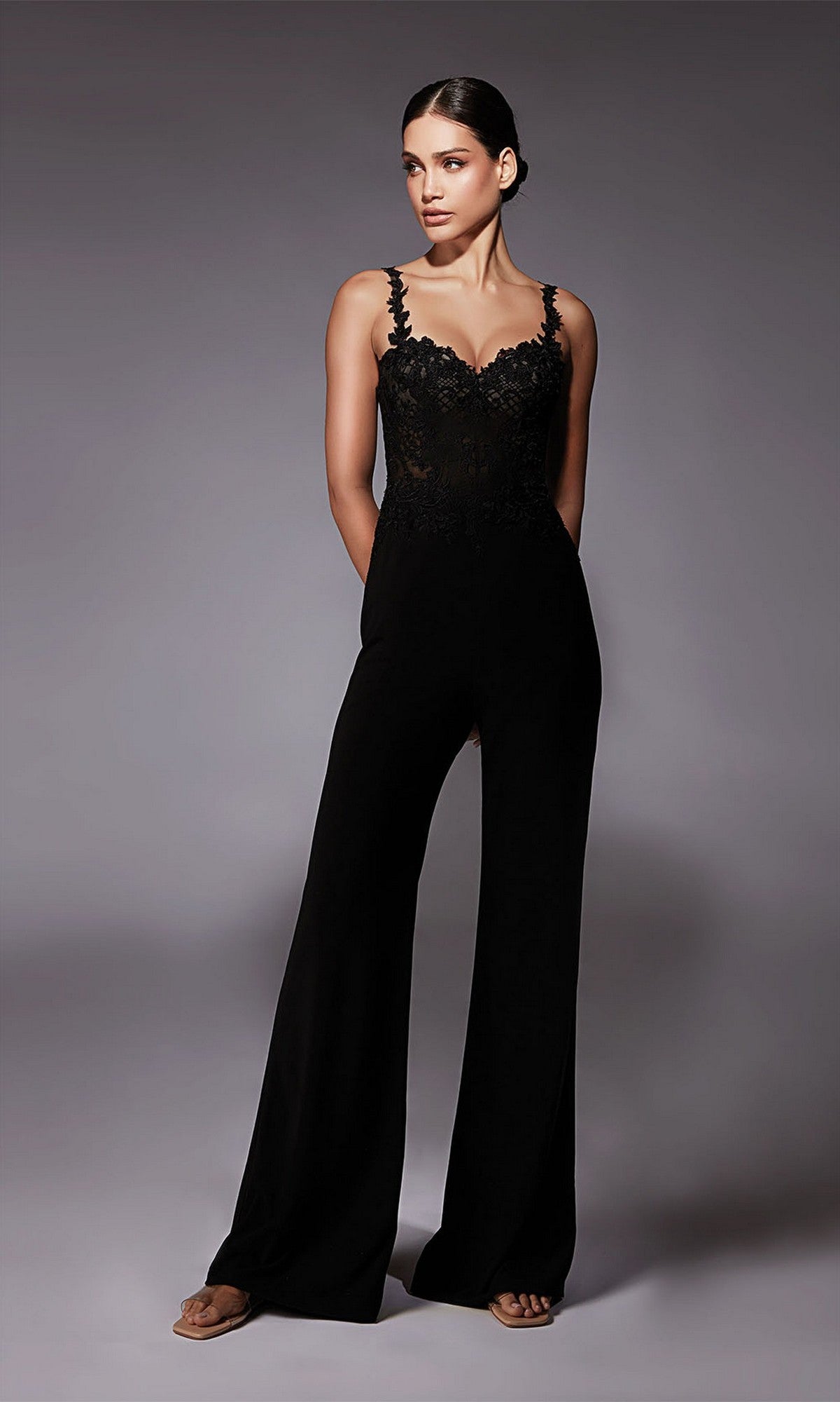 Woman wearing the Long Formal Jumpsuit: Alyce 70074, featuring lace trim and wide legs, stands against a plain gray background with her hands in her pockets.